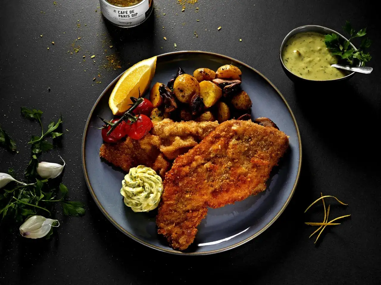 Ein knuspriges Wiener Schnitzel mit Bratkartoffeln, Tomaten, Zitrone und Kr&auml;uterbutter. Dazu eine cremige Sauce.