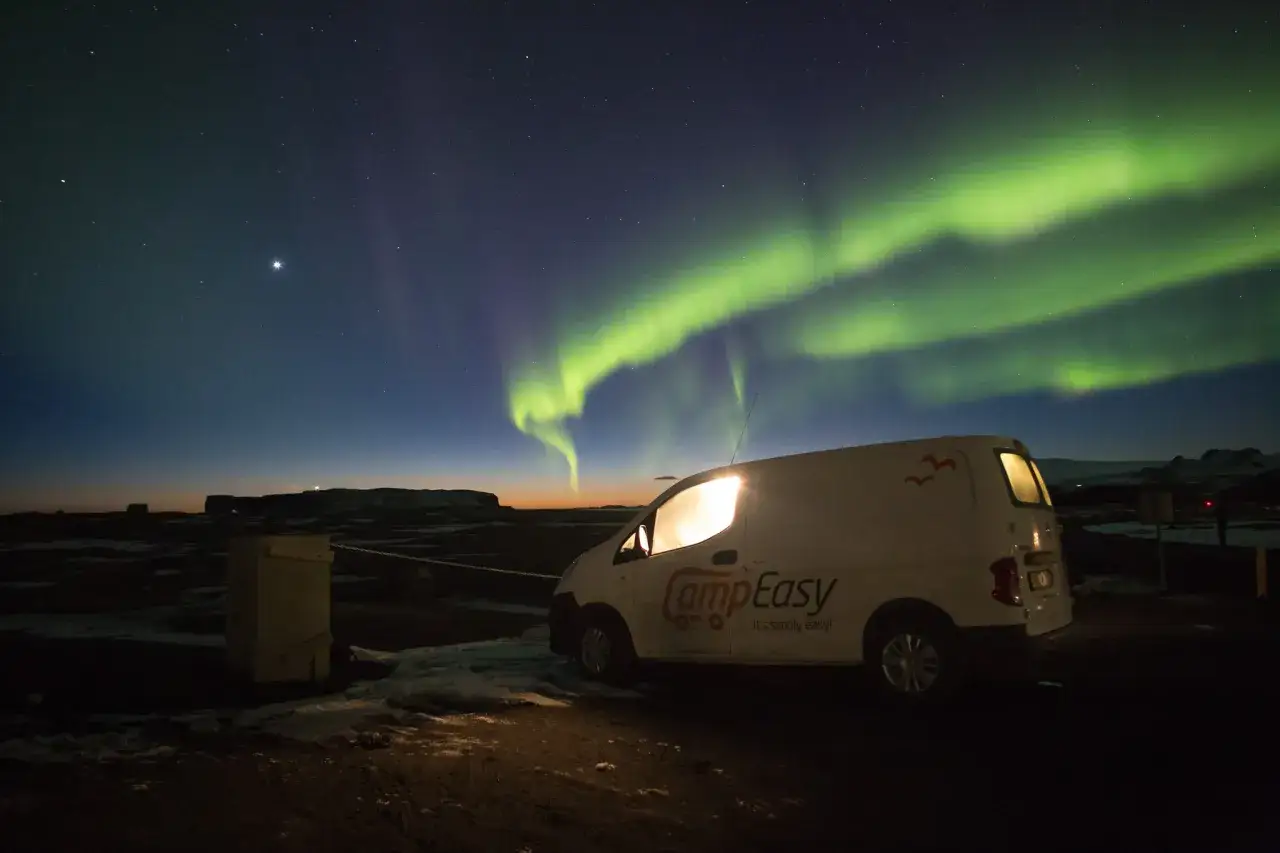 Ein Campervan steht unter dem magischen Licht der Aurora Borealis. Gr&uuml;ne Schleier tanzen am sternenklaren Himmel &uuml;ber einer dunklen Landschaft.