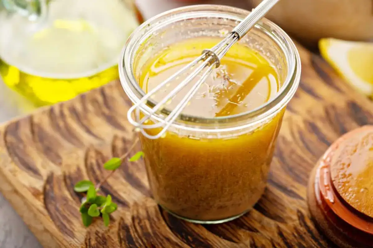 Ein Glas mit selbstgemachtem Salat Dressing, darin ein kleiner Schneebesen. Daneben ein paar Kr&auml;uter und eine Zitronenspalte.