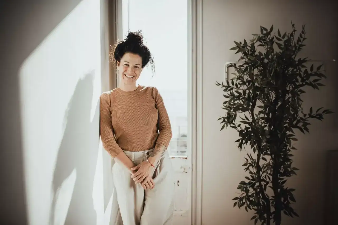 Mujer sonriente apoya en pared, con planta al lado. Sus **youtooproject opiniones** son positivas, reflejando su felicidad.