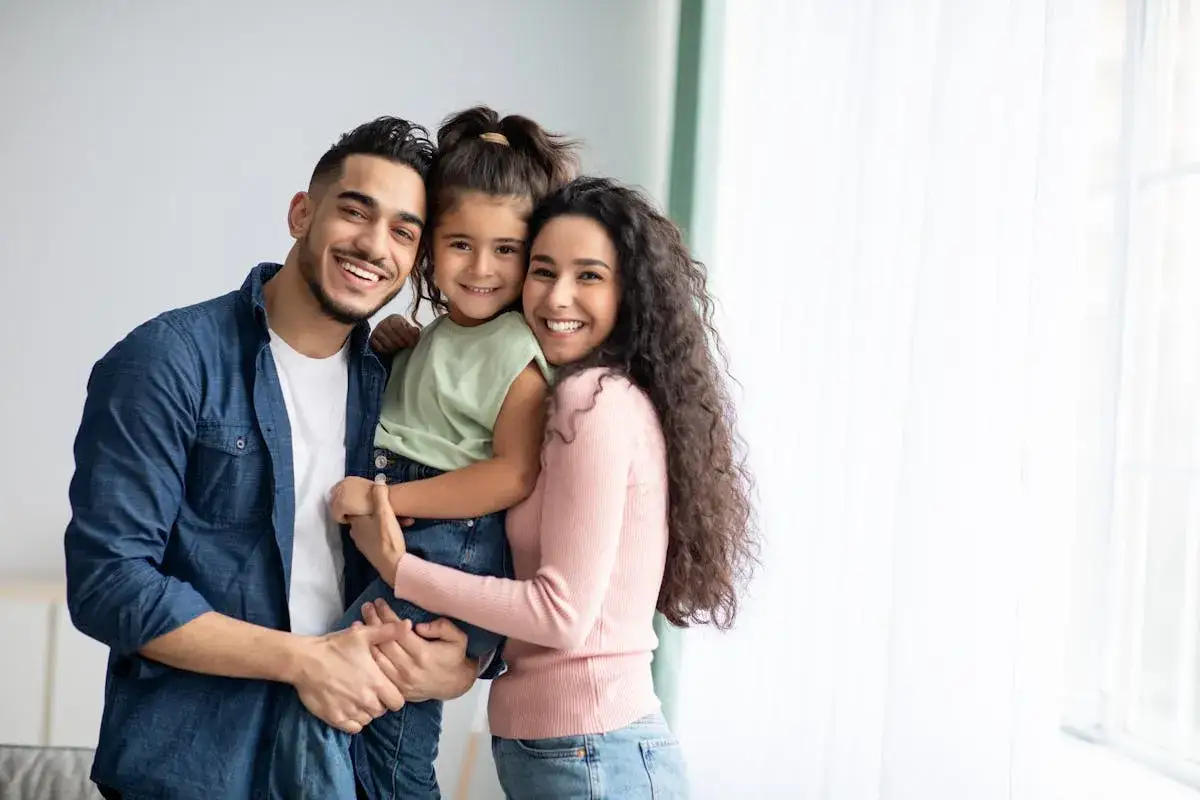 Un couple souriant et leur fille, une famille avec un nom de famille tunisien, posent joyeusement devant une fen&ecirc;tre.