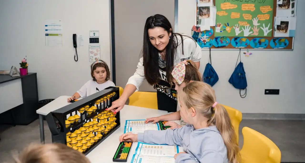 Nauczycielka pokazuje dzieciom liczydło i inne przedmioty szkolne. Dziewczynki uczą się matematyki.