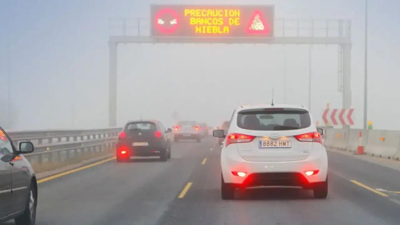 Tr&aacute;fico denso en carretera con niebla. Los coches encienden sus luces antiniebla para mejorar la visibilidad. Se&ntilde;al de advertencia de bancos de niebla.