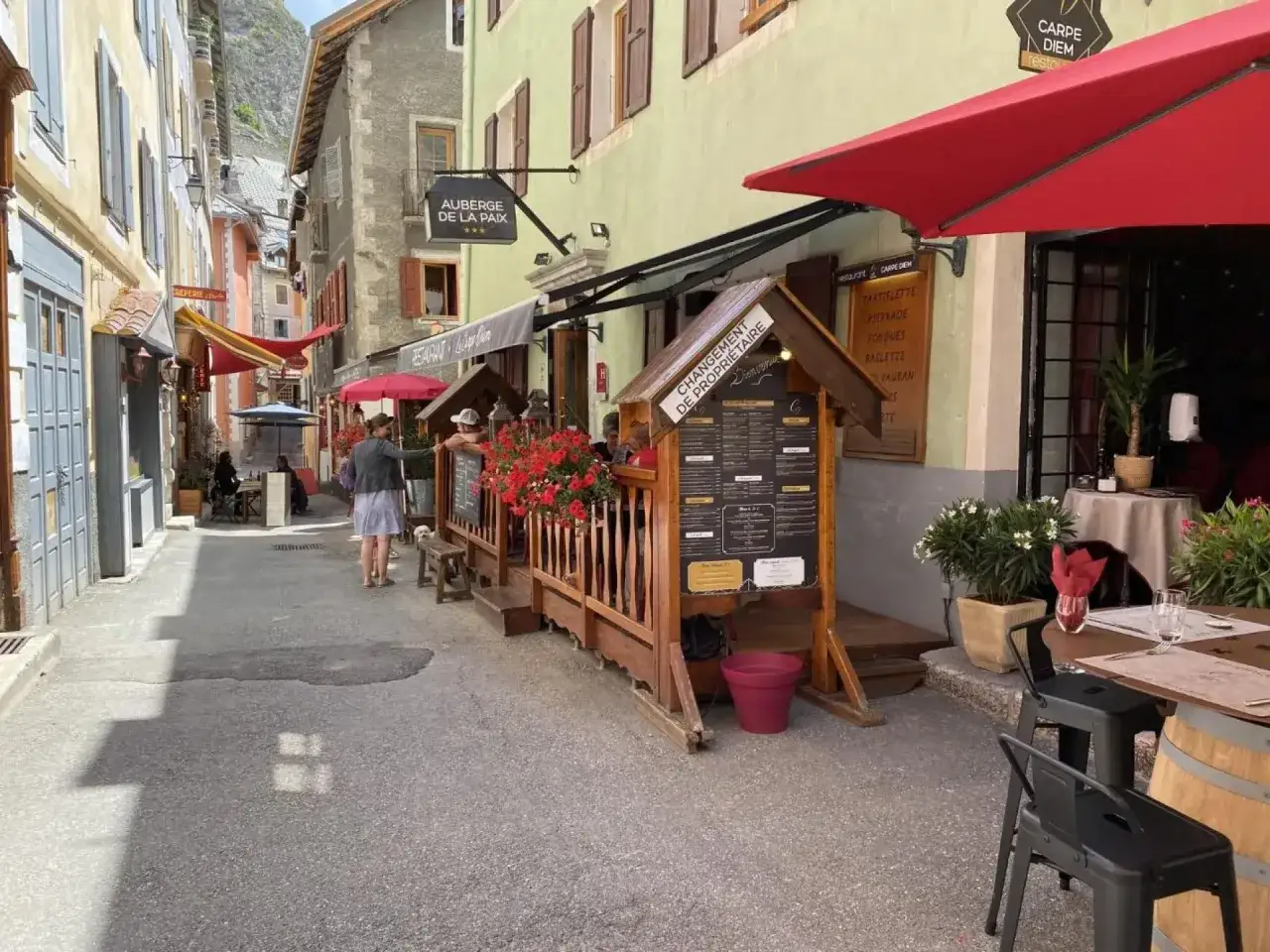 Rue animée de Briançon avec des restaurants et terrasses fleuries. L'Auberge de la Paix et le restaurant Carpe Diem invitent à la découverte des spécialités locales.