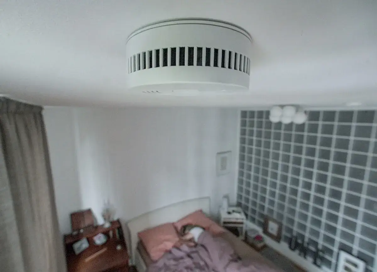 Ein weißer feuermelder wohnung ist an der Decke montiert. Darunter ein Bett mit rosa Kissen und Decken, daneben ein Nachttisch und ein Schreibtisch.