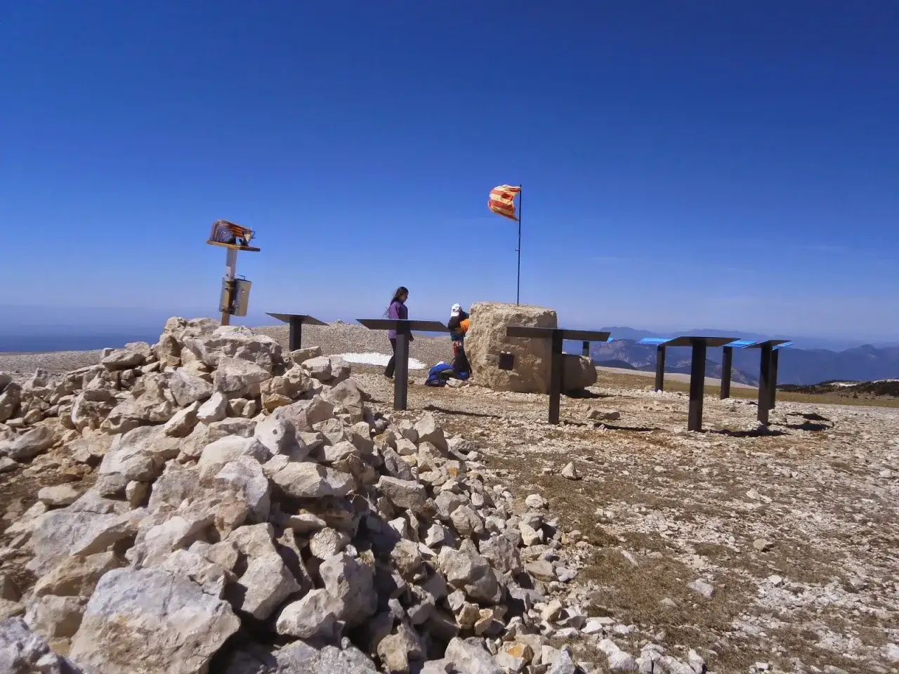 En el pedró dels quatre batlles, dos personas observan el paisaje montañoso bajo un cielo azul. Una bandera ondea al viento.
