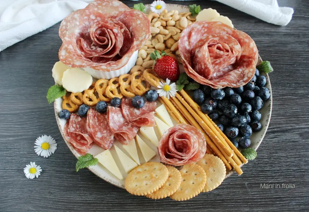 Un invitante tagliere di antipasti con salumi a forma di rosa, formaggio, fragole, mirtilli, pretzel e cracker.