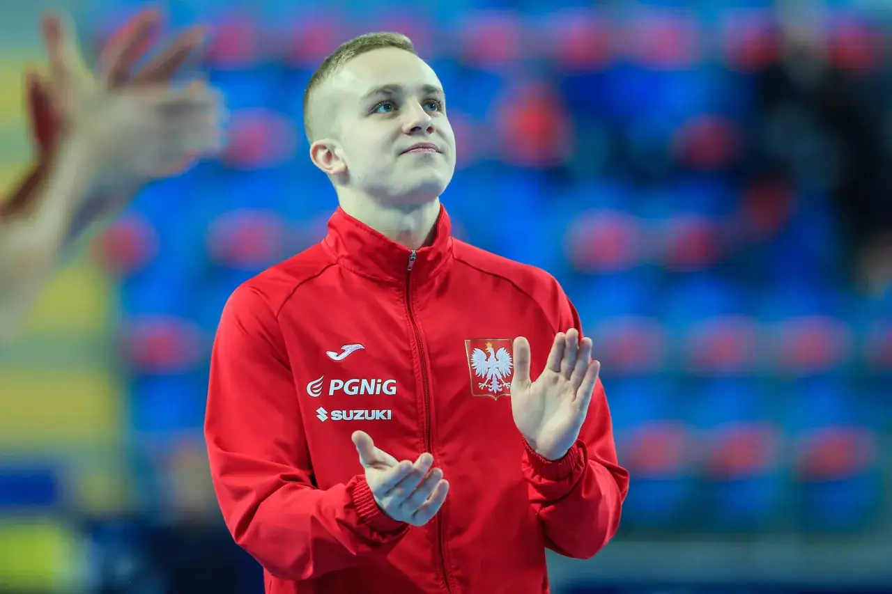 Adam Jędraszek w czerwonej kurtce z orłem na piersi, z dłońmi uniesionymi w geście radości.