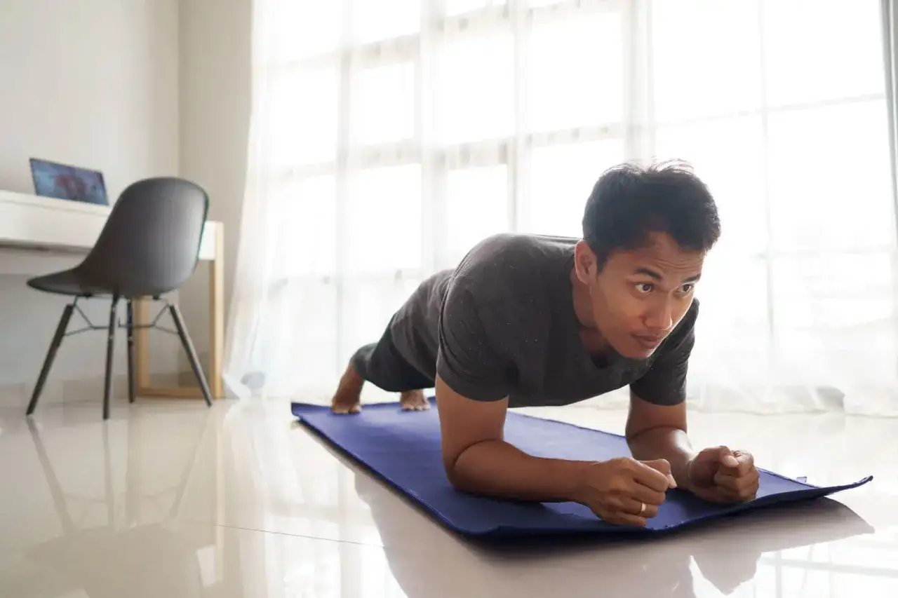 Un homme fait un exercice de gainage sur un tapis bleu, profitant du sport à la maison près d'une grande fenêtre lumineuse.