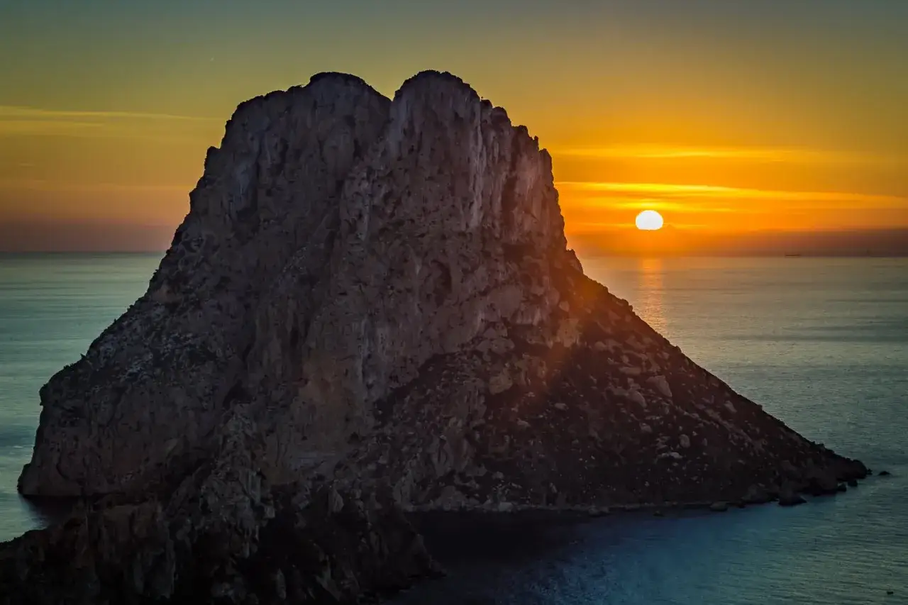El sol se pone detrás del imponente Ojo de Es Vedrà, tiñendo el mar de tonos dorados y anaranjados.