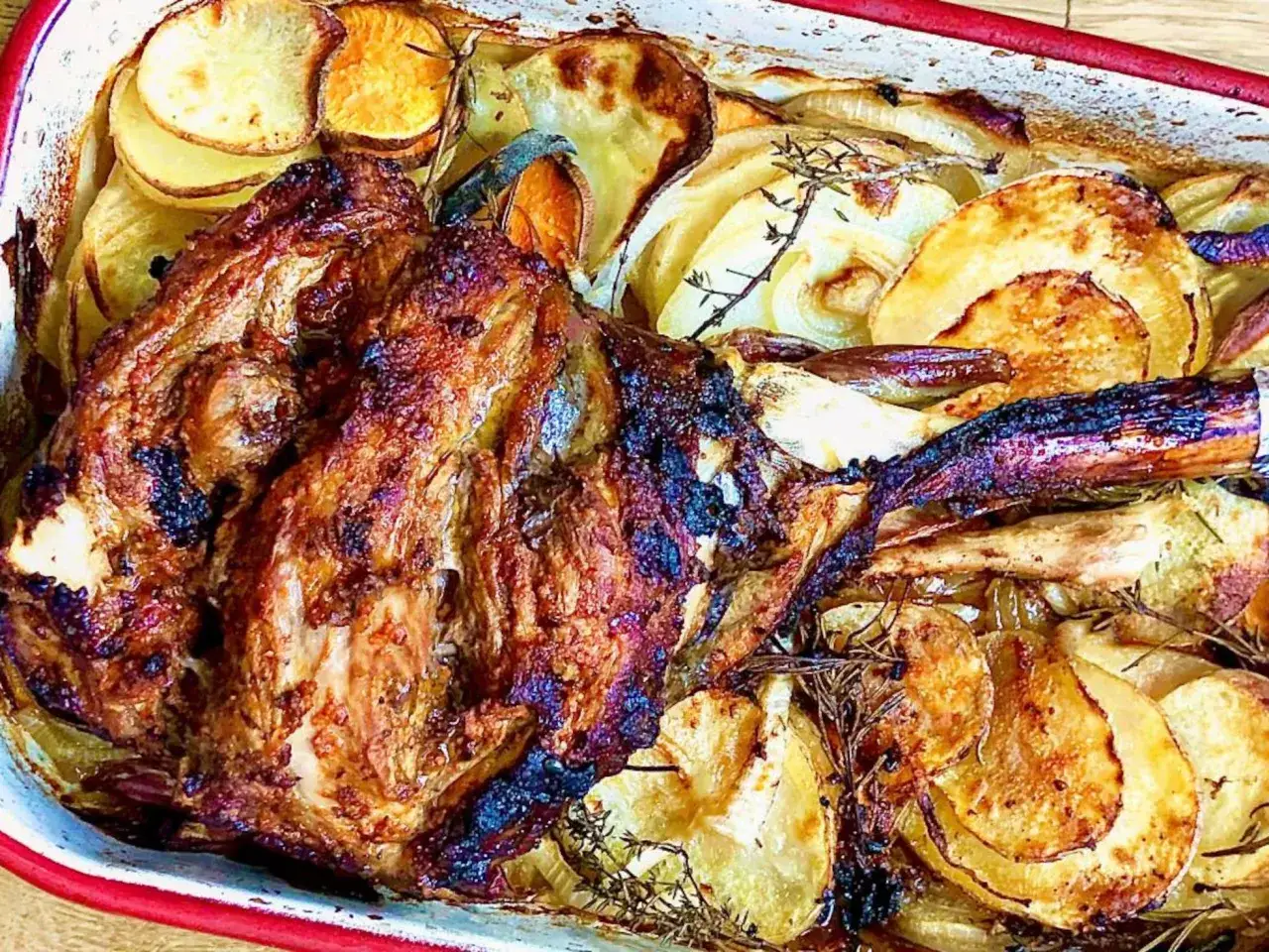 Pierna de cordero asada, dorada y jugosa, acompañada de patatas y boniatos en rodajas, todo horneado con hierbas aromáticas.