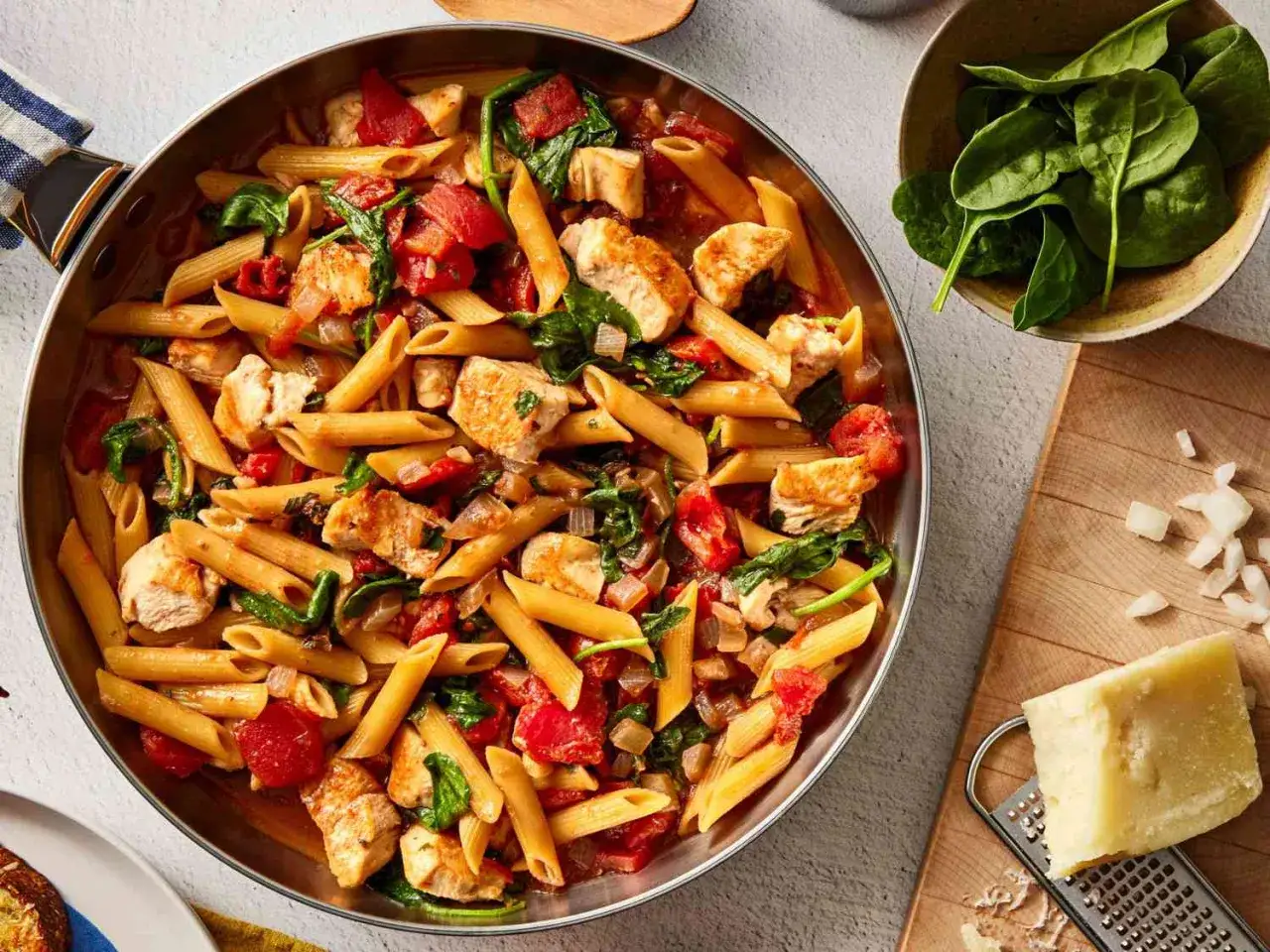 Penne z kurczakiem i szpinakiem w sosie pomidorowym, podane w patelni. Obok miseczka świeżego szpinaku i kawałek parmezanu.