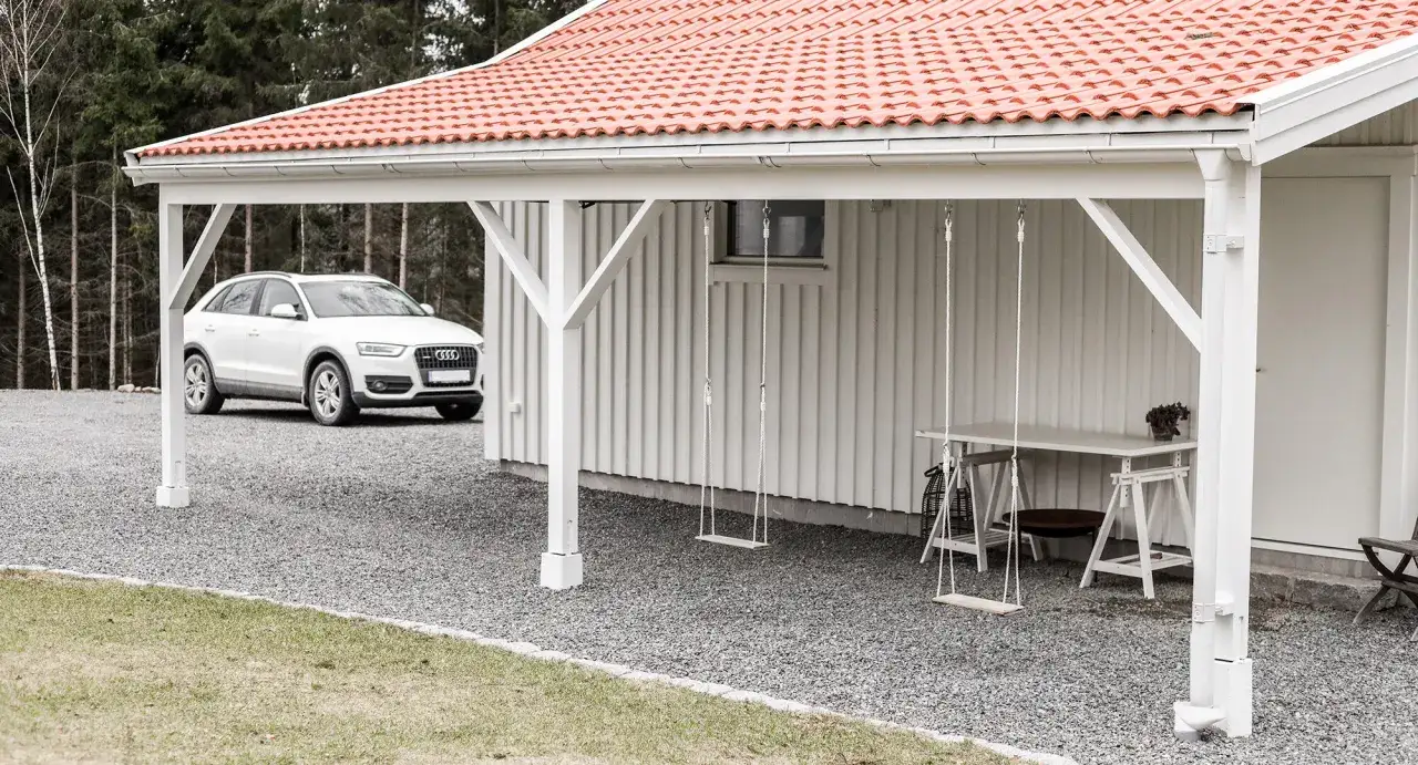 Vit bil parkerad vid ett hus med utbyggnad p&aring; l&aring;ngsidan. Gungor och bord under taket.