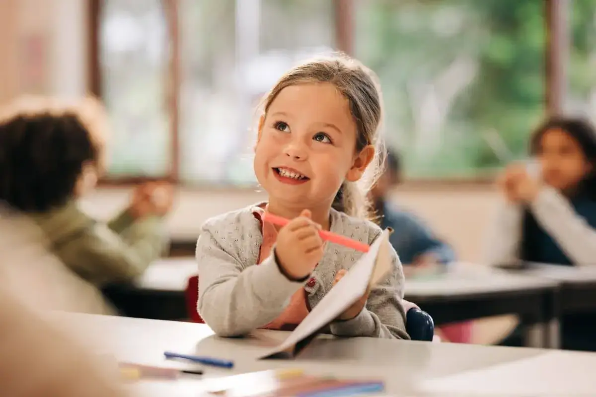 Ni&ntilde;a sonriente en clase, con un l&aacute;piz rosa en la mano, lista para su iniciaci&oacute;n a la lectura.