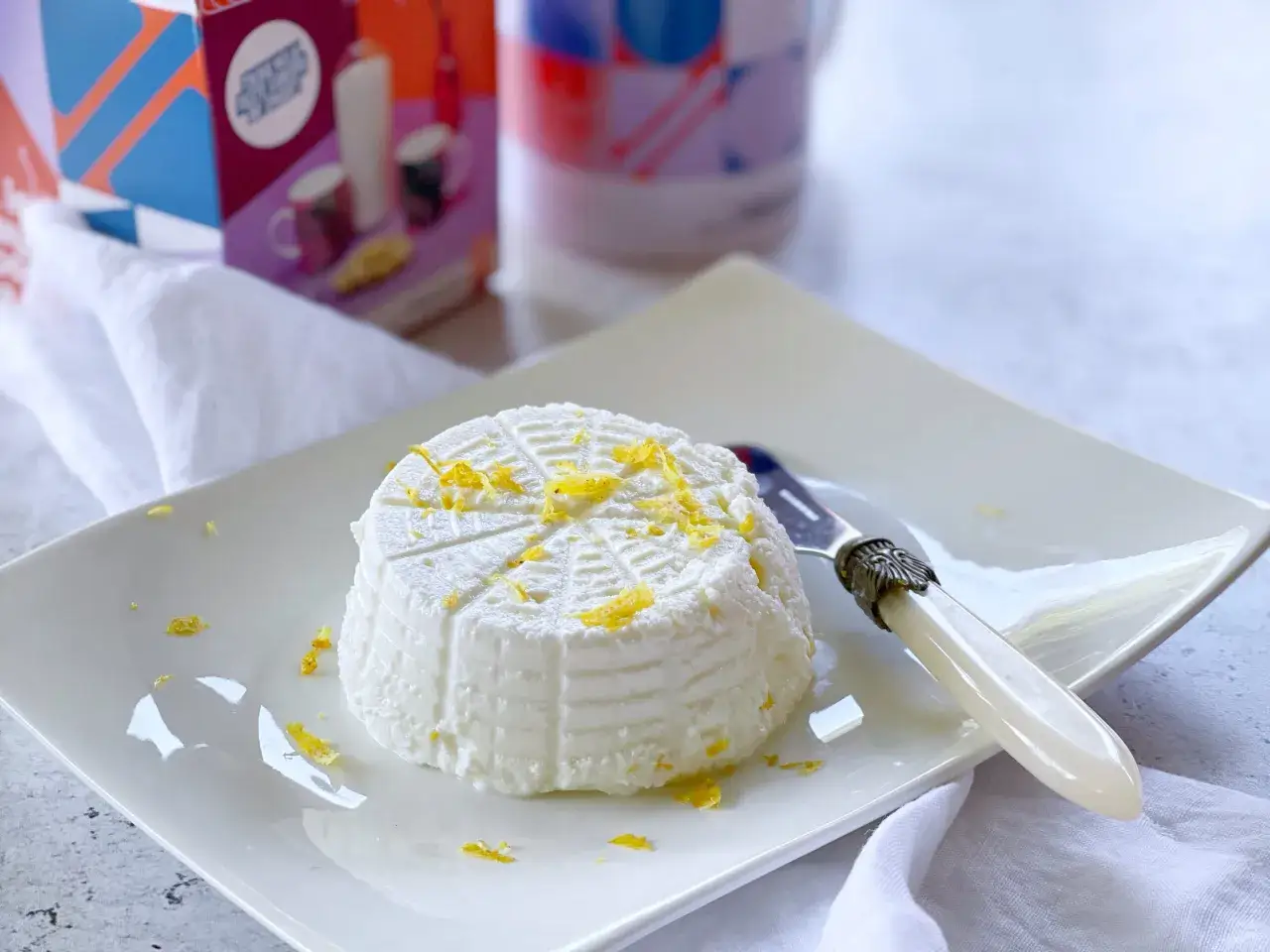 Ricotta fatta in casa, fresca e profumata, guarnita con scorza di limone su un piatto bianco. Un cucchiaino decorato &egrave; pronto per assaggiarla.