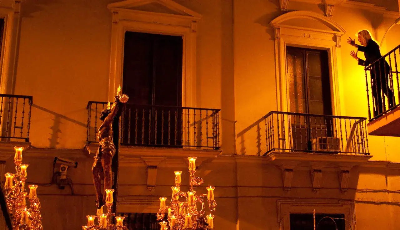 Procesi&oacute;n nocturna con Cristo crucificado, candelabros y una mujer en un balc&oacute;n. El **saeta significado** se siente en la devoci&oacute;n.