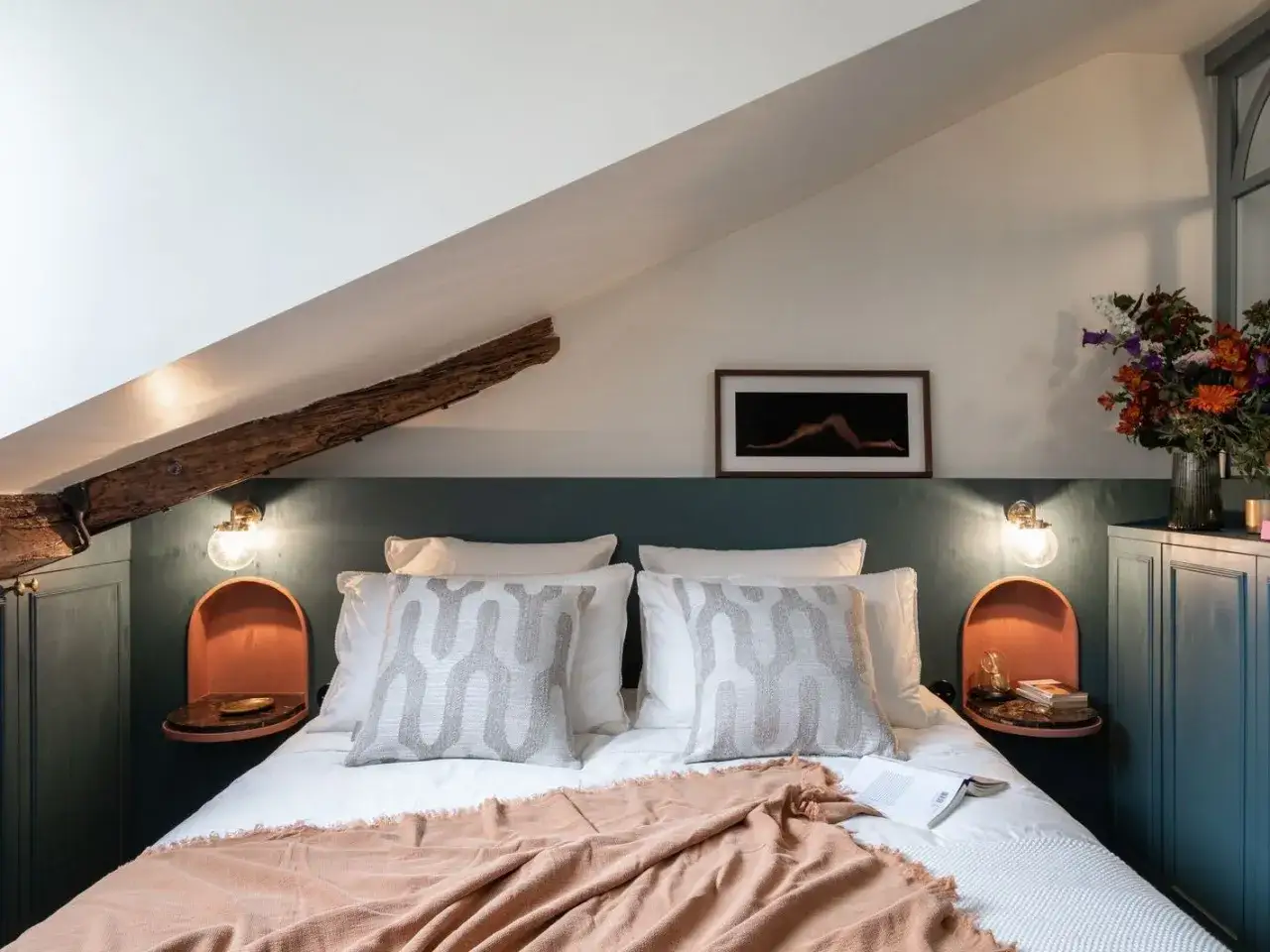 Tête de lit sous pente avec poutre apparente, coussins graphiques et tableau artistique. Ambiance cosy et moderne.