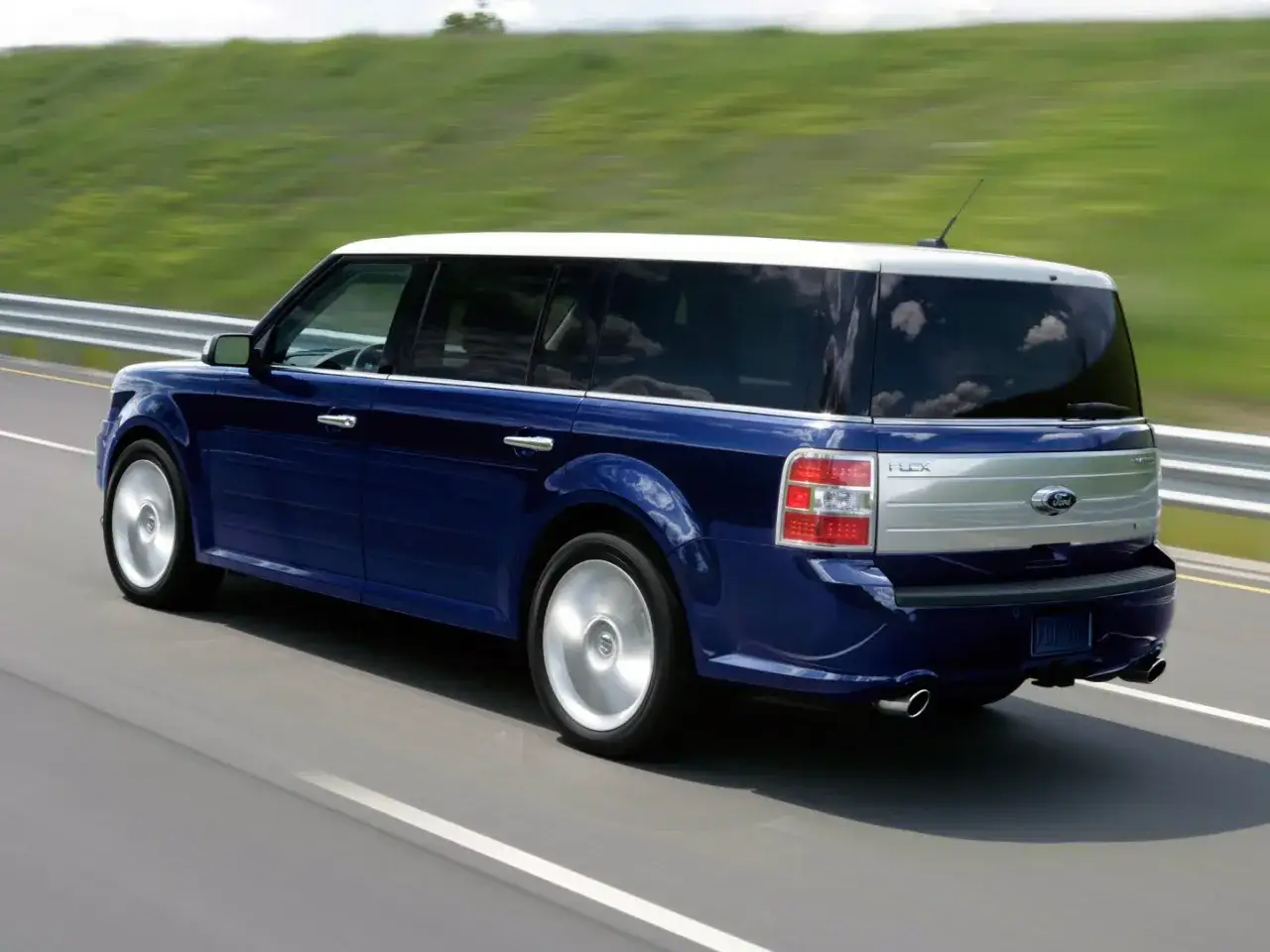 Niebieski Ford Flex z białym dachem pędzi po drodze, otoczony zielonymi wzgórzami.