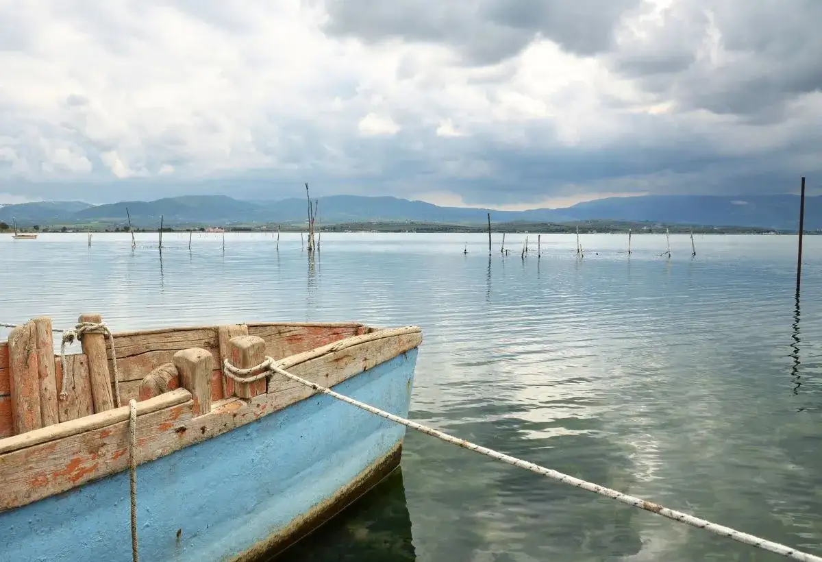 Barca blu ormeggiata in un lago tranquillo, con pali di legno che emergono dall'acqua e montagne sullo sfondo. Un'atmosfera serena a Cagnano Varano.