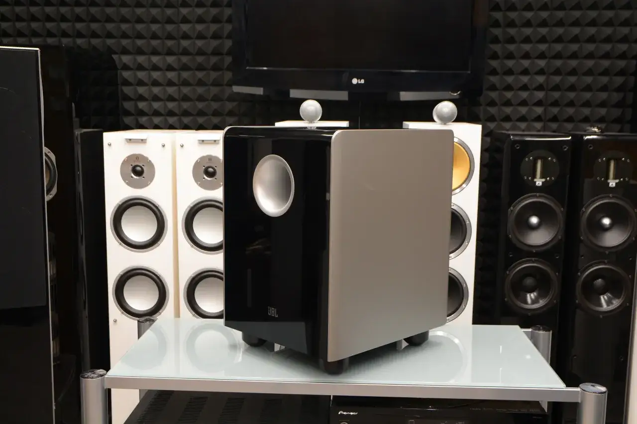 Czarny, błyszczący jbl subwoofer aktywny estradowy z szarym panelem, otoczony innymi głośnikami.