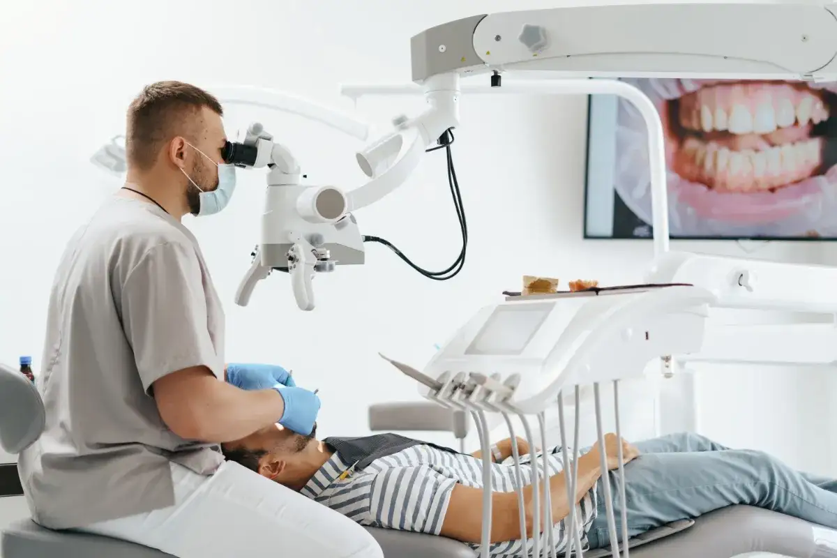 Dentysta w masce i rękawiczkach bada pacjenta pod mikroskopem. Na ekranie w tle widać zbliżenie uśmiechu. To nowoczesna mta stomatologia.