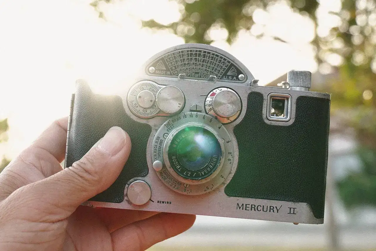 A hand holds a vintage Mercury II camera, a classic piece of photography history, with the sun glinting off its lens.