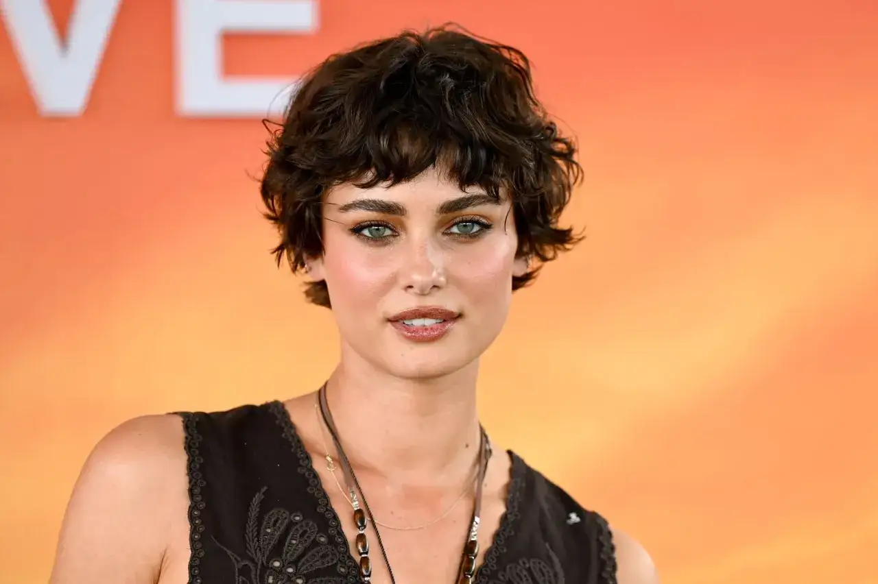 Mujer con peinados pelo corto ondulado, mirada intensa y labios brillantes, luciendo un chaleco oscuro.