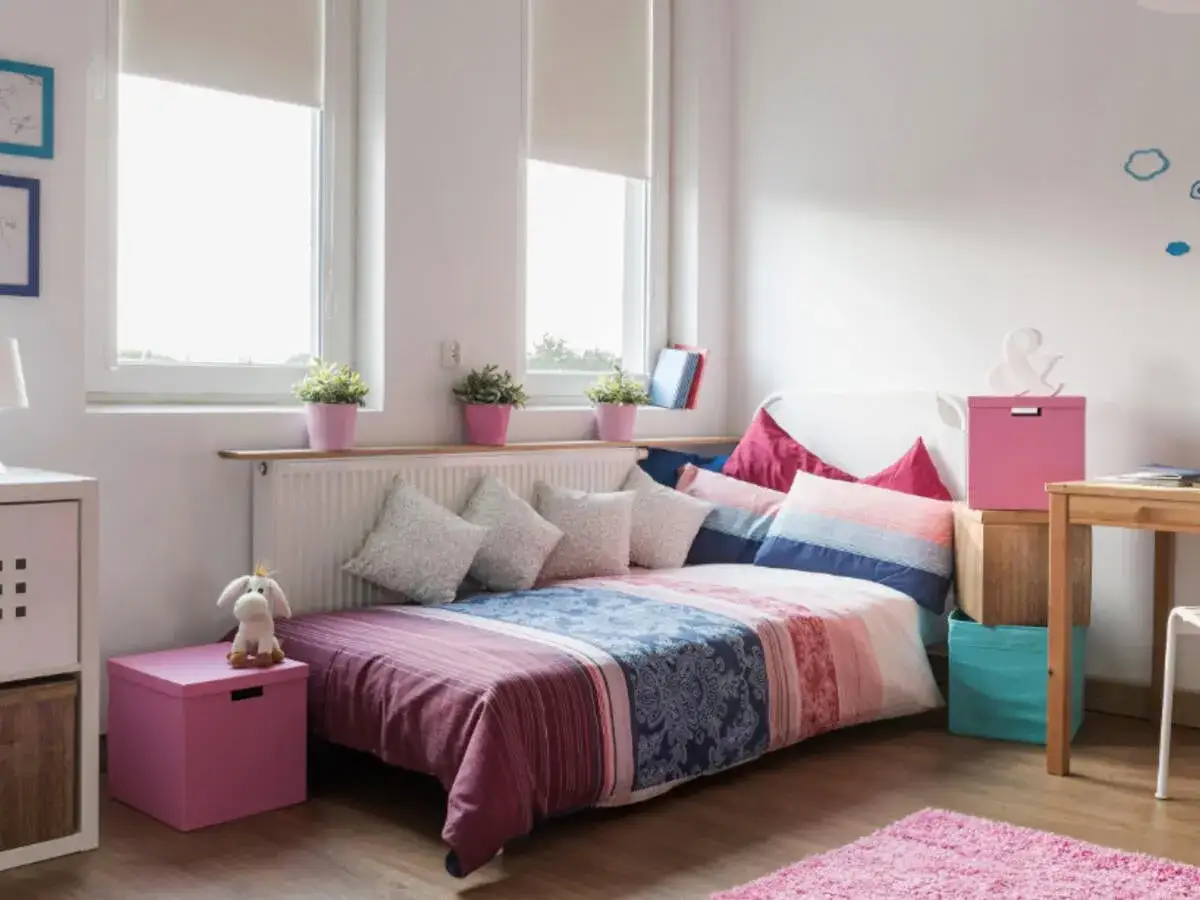 Ein gem&uuml;tliches kleine jugendzimmer mit einem Bett, das mit bunten Kissen und Decken dekoriert ist. Pflanzen auf der Fensterbank und ein Teddyb&auml;r auf einer rosa Kiste sorgen f&uuml;r Gem&uuml;tlichkeit.
