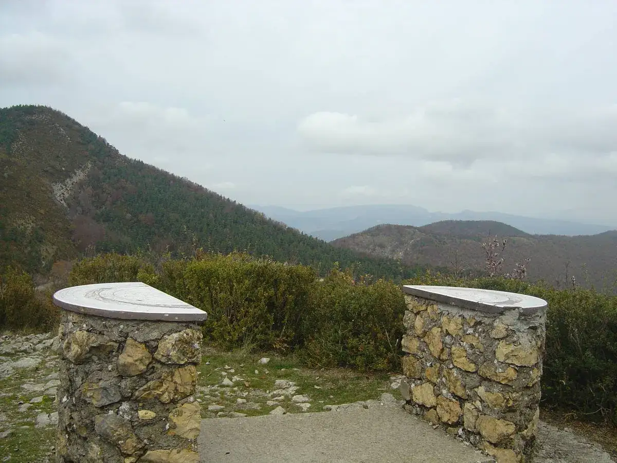 Vue panoramique depuis le col de Perty, avec des piliers en pierre et des tables d'orientation. Paysage montagneux couvert de forêts.