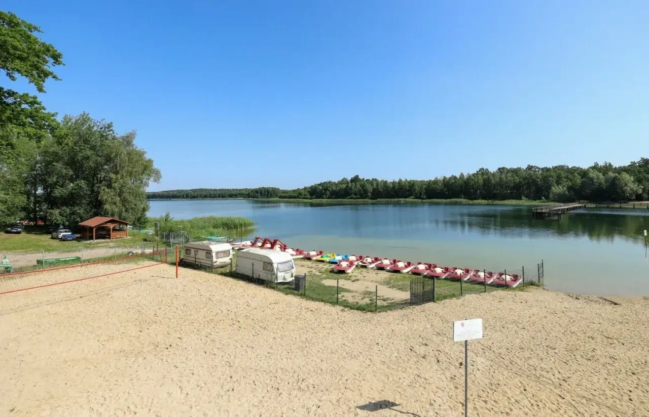Piękny dzień nad jeziorem Kamionki. Piaskowa plaża, siatkówka, przyczepy kempingowe i rzędy łódek czekają na gości.