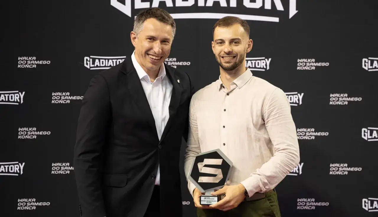Paweł Paterek z nagrodą w kształcie litery S, uśmiechnięty obok mężczyzny w garniturze na tle ścianki z napisem