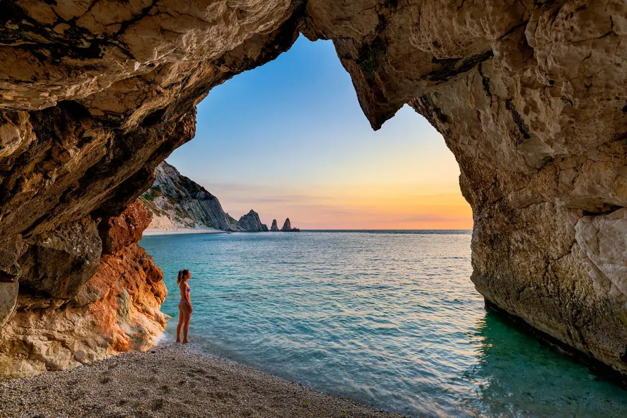 Una ragazza ammira il tramonto sul mare nelle Marche, vista da una grotta. Le rocce scolpite dal mare creano una cornice naturale.