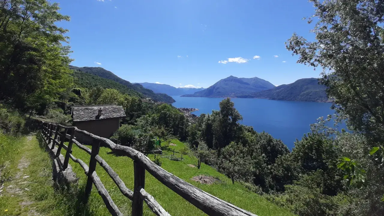 Sentiero viandante con vista su lago e montagne. Una piccola casa in pietra e una staccionata in legno accompagnano il cammino.