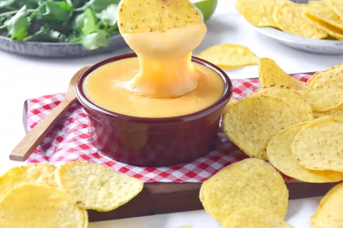 Un totopo se sumerge en una cremosa salsa cheddar, rodeado de m&aacute;s totopos y cilantro fresco.