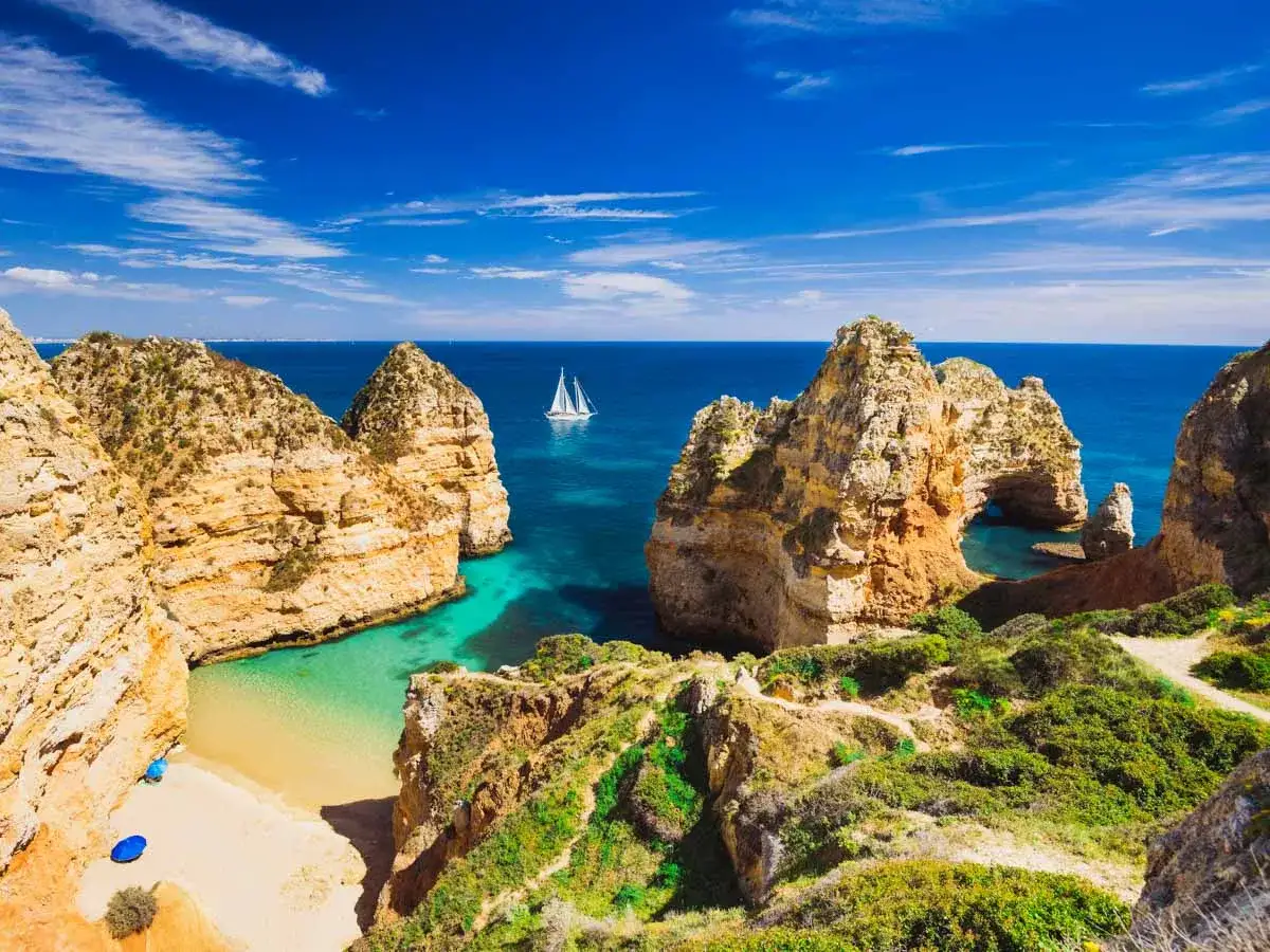 Traumhafte Felsformationen und eine kleine Bucht an der Costa Verde, Portugal. Ein Segelboot gleitet auf dem türkisfarbenen Meer.