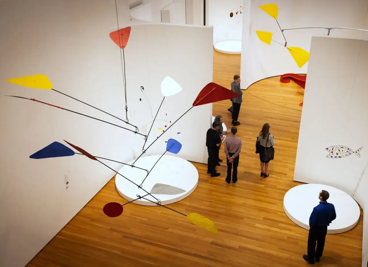 Un mobile Calder aux formes g&eacute;om&eacute;triques color&eacute;es flotte dans un mus&eacute;e, admir&eacute; par des visiteurs.