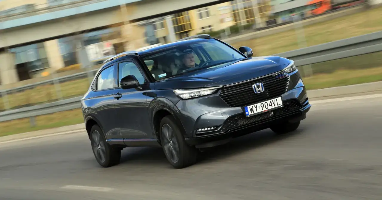 Ciemnoszary Honda HR-V jedzie po drodze, z rozmytym tłem budynków i drzew.