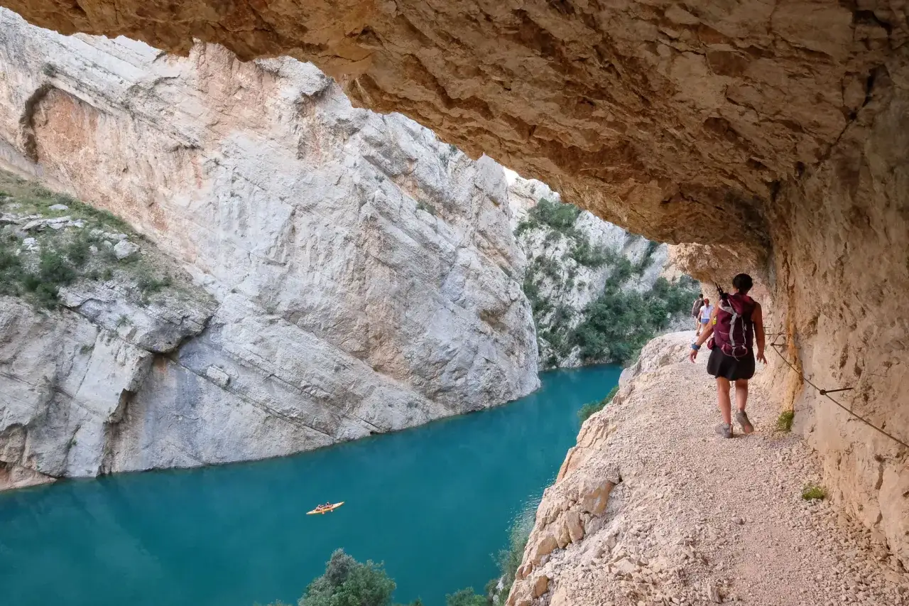 Senderismo por un ca&ntilde;&oacute;n con un r&iacute;o turquesa. Personas caminan por un sendero estrecho bajo una roca. Kayak en el agua. Ideales excursiones cerca de Tremp.
