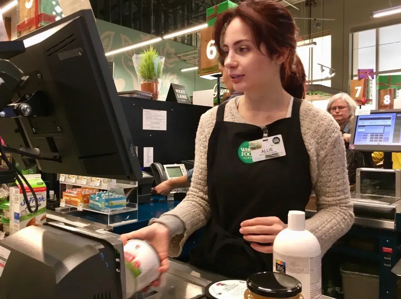 Cassiera al supermercato, Allie, con il suo badge, mentre scansiona un prodotto. Il suo stipendio da cassiera supermercato è meritato.