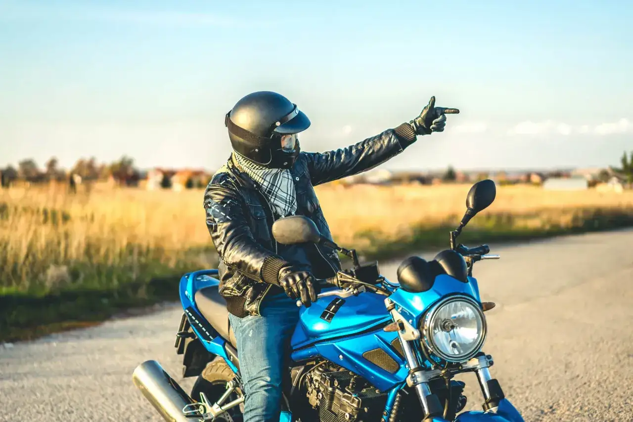Motocyklista w kasku i sk&oacute;rzanej kurtce wskazuje drogę. W tle złote pola i błękitne niebo. To znaki motocyklist&oacute;w, kt&oacute;re łączą wolność i przygodę.