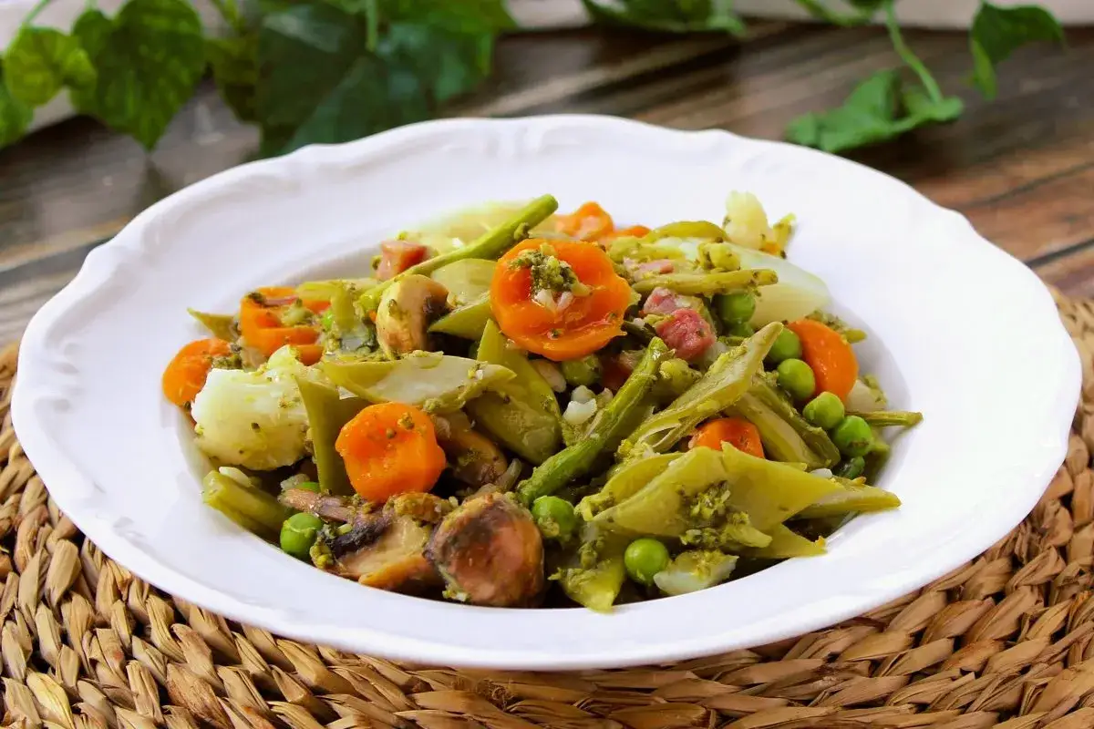 Un plato de menestra de verduras con patatas, zanahorias, judías verdes, guisantes y champiñones, todo aderezado con una salsa verde.