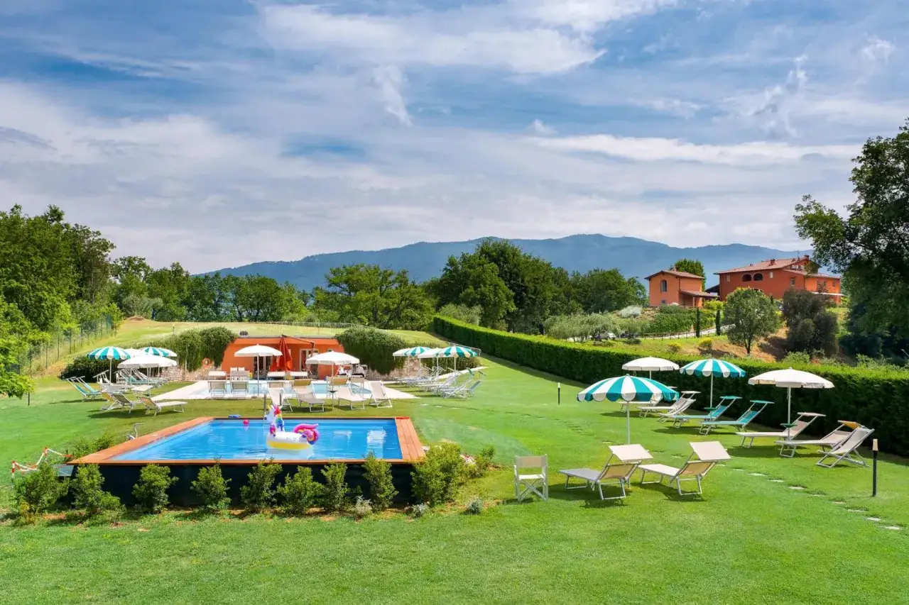 Un'oasi di pace in **Toscana con neonato**: piscina, lettini e ombrelloni su un prato verde, con case coloniche e colline sullo sfondo.