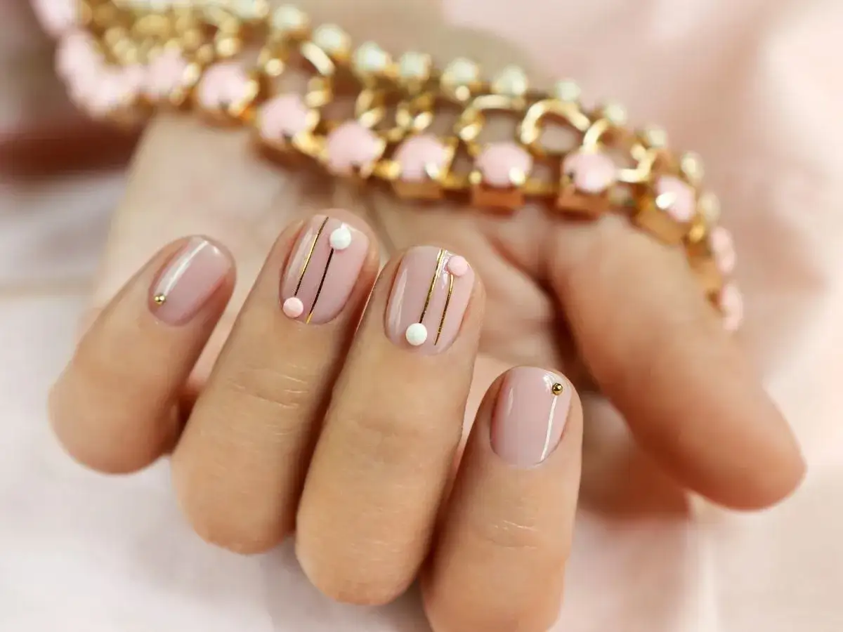 Delikatny różowy manicure z ozdobami: złote linie, koraliki i kuleczki na krótkiej i szerokiej płytce paznokcia.