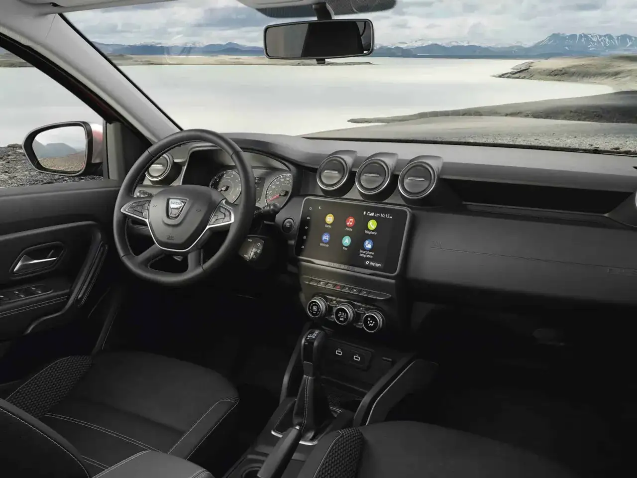 Wnętrze nowej Dacii Duster z ekranem dotykowym, kierownicą i widokiem na jezioro i góry.