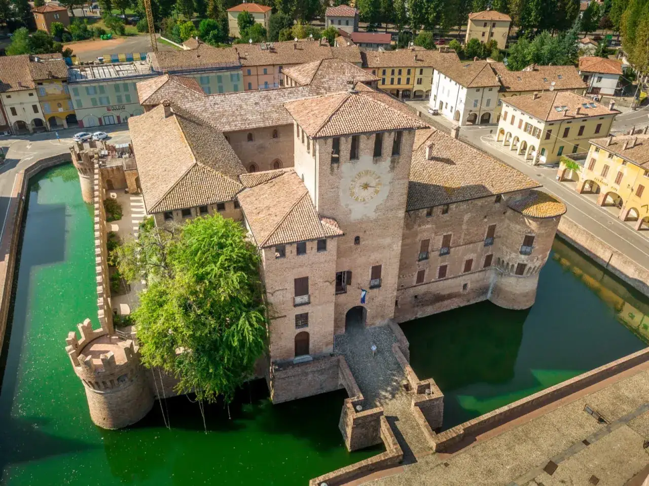 Antico monumento in Emilia Romagna: un castello medievale circondato da un fossato verde smeraldo, con torri e un orologio sulla torre principale.