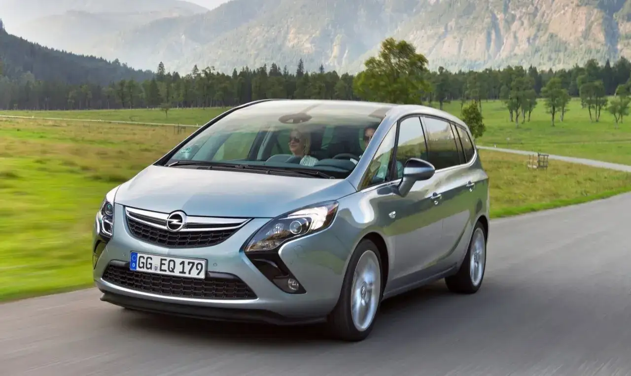 Silberner Opel Zafira C fährt auf einer Landstraße durch eine grüne Landschaft mit Bergen im Hintergrund.