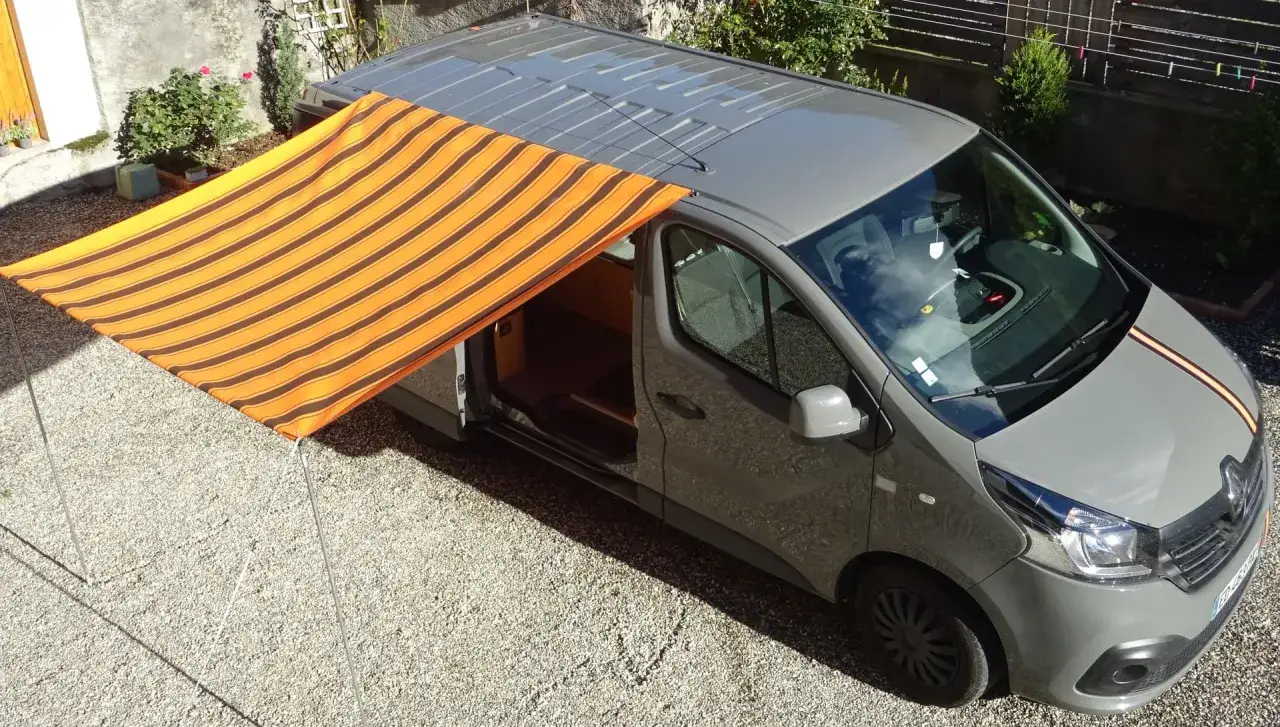 Un store banne rayé orange et marron est installé sur le côté d'un fourgon gris, offrant un abri ombragé.