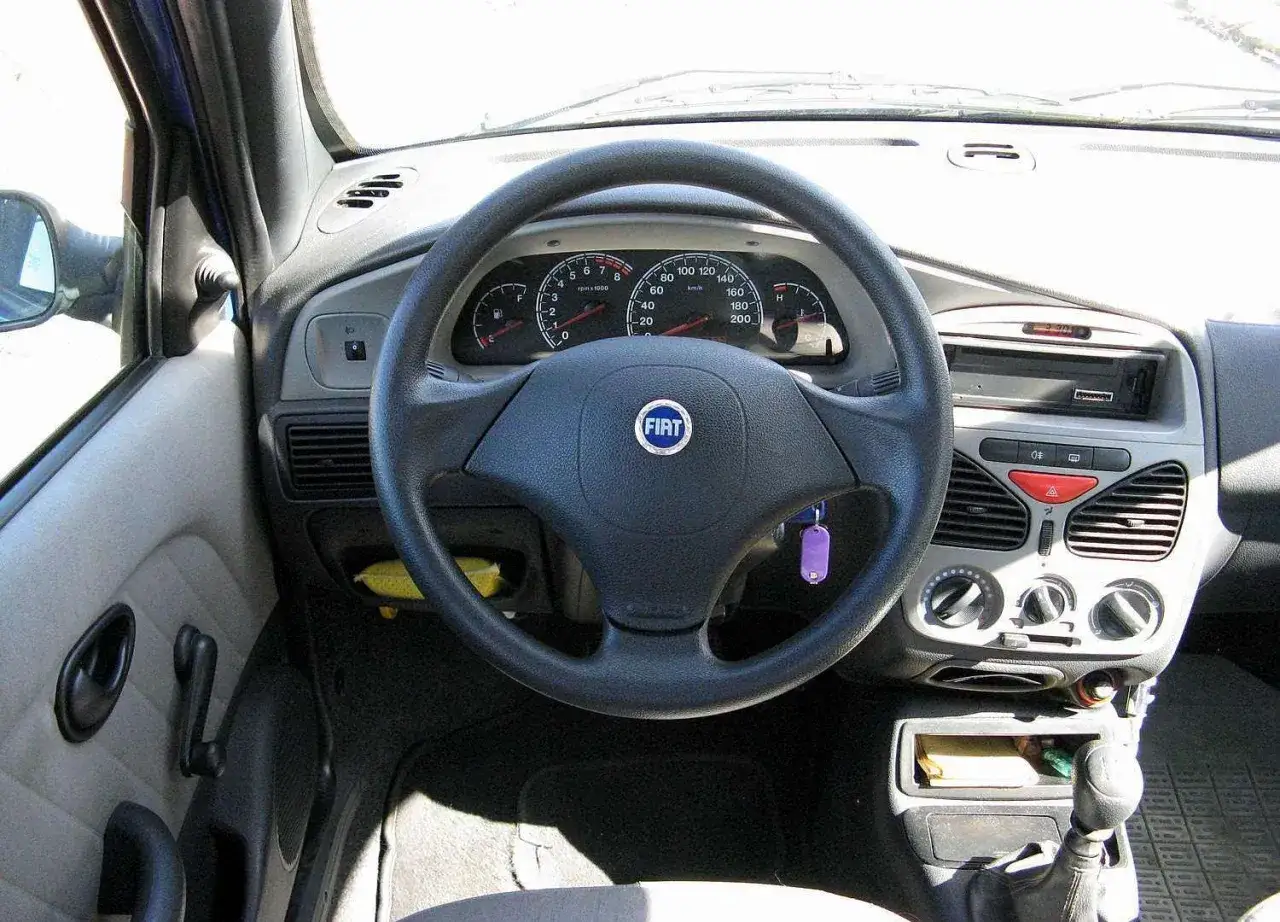 Wnętrze Fiata Albea: kierownica z logo FIAT, deska rozdzielcza z zegarami i radiem, widoczna klamka drzwi.
