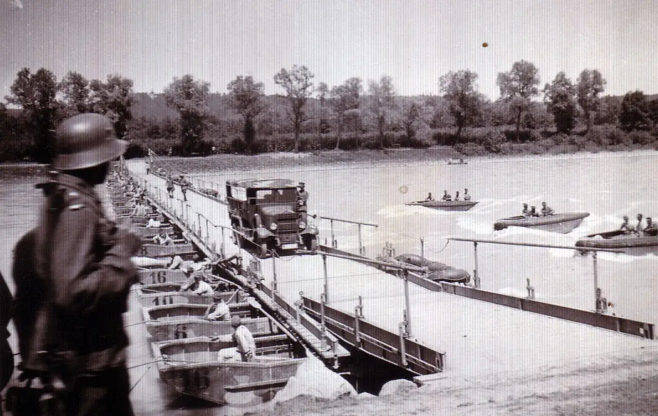 Ein deutscher Kfz-Pionier überwacht den Aufbau einer Pontonbrücke über einen Fluss. Boote mit Soldaten kreuzen auf dem Wasser.