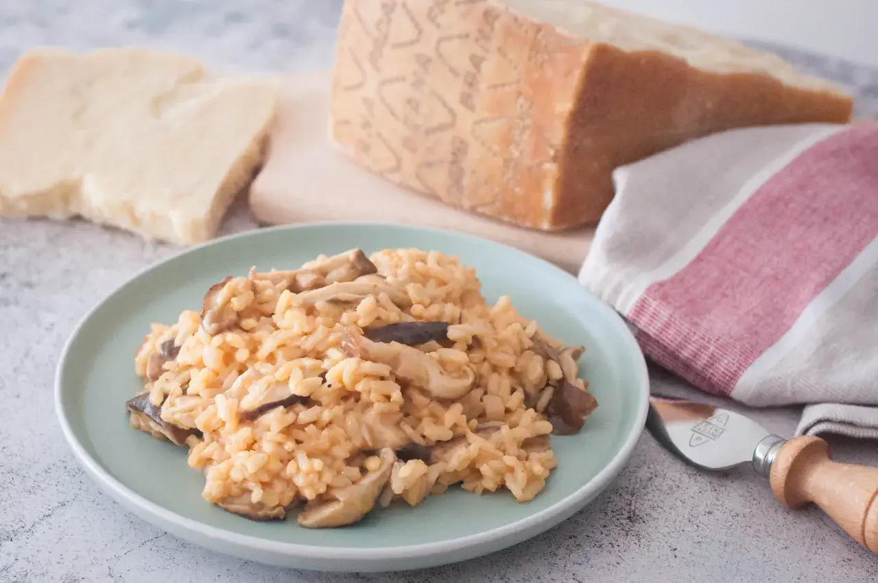 Un cremoso risotto de setas servido en un plato verde, con trozos de queso Parmesano y un pa&ntilde;o a rayas al lado.